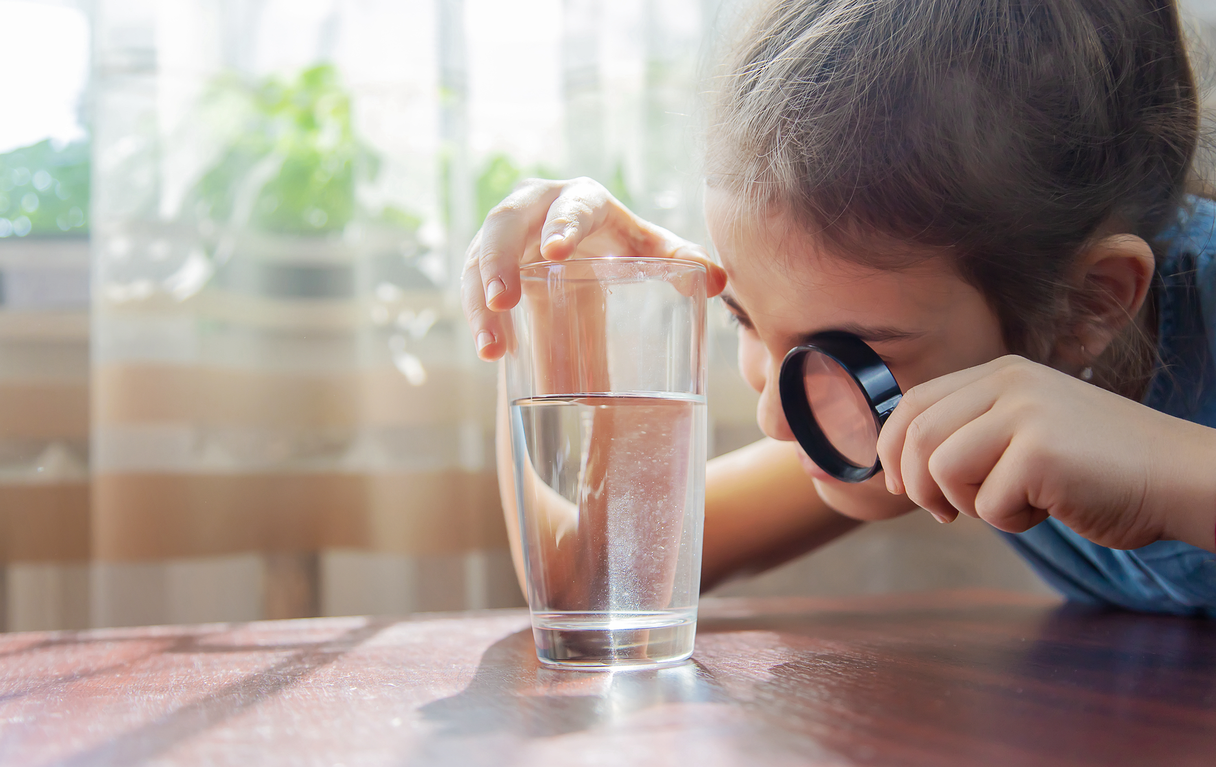 Une jeune fille examine un verre d’eau du robinet avec une loupe, illustrant la problématique des substances cancérogènes présentes dans l'eau potable.